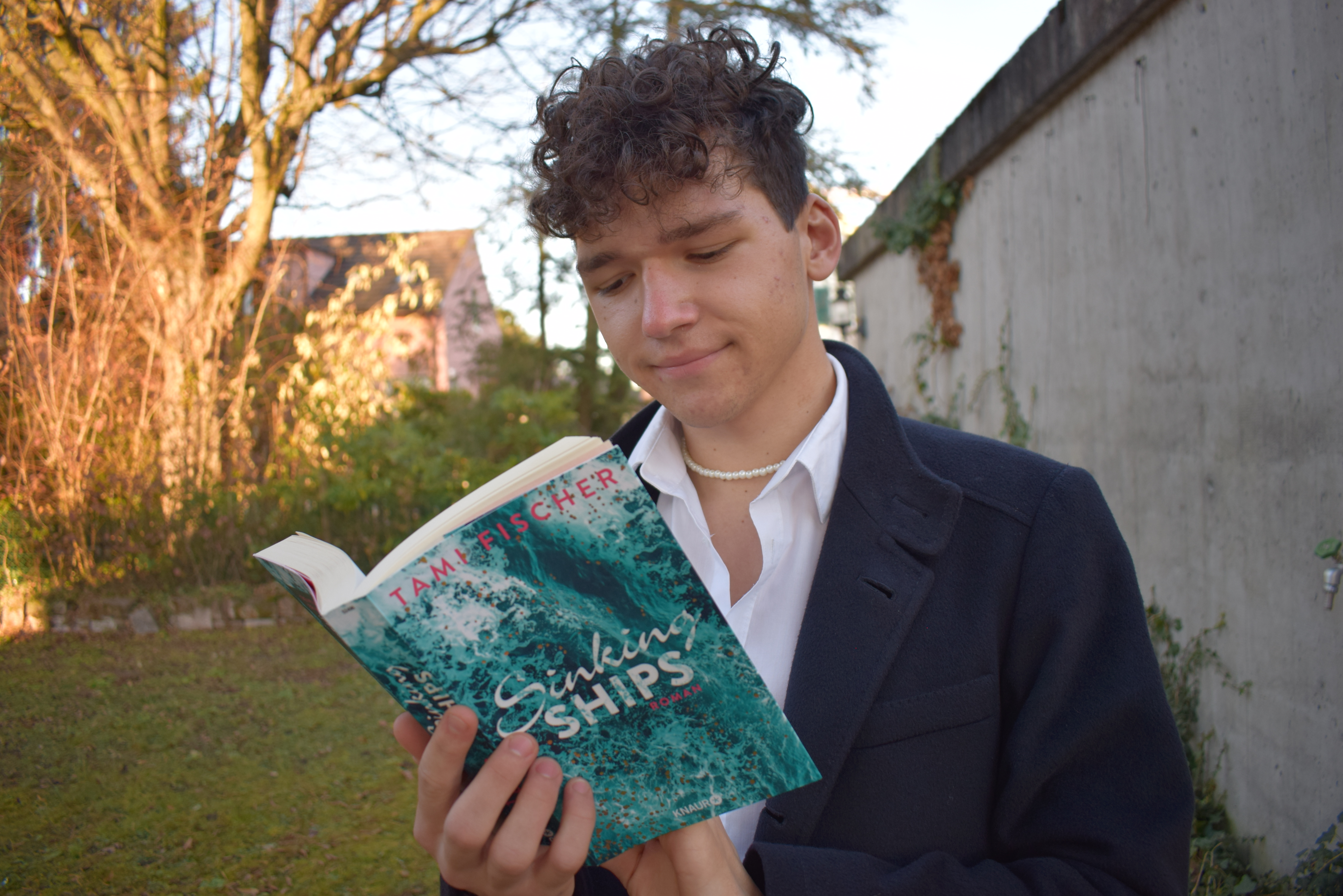 Sinking Ships Rezension Tami Fischer Josia Jourdan - Junger Mann im Hemd, mit Buch und Perlenkette im Garten - lesender junger Mann