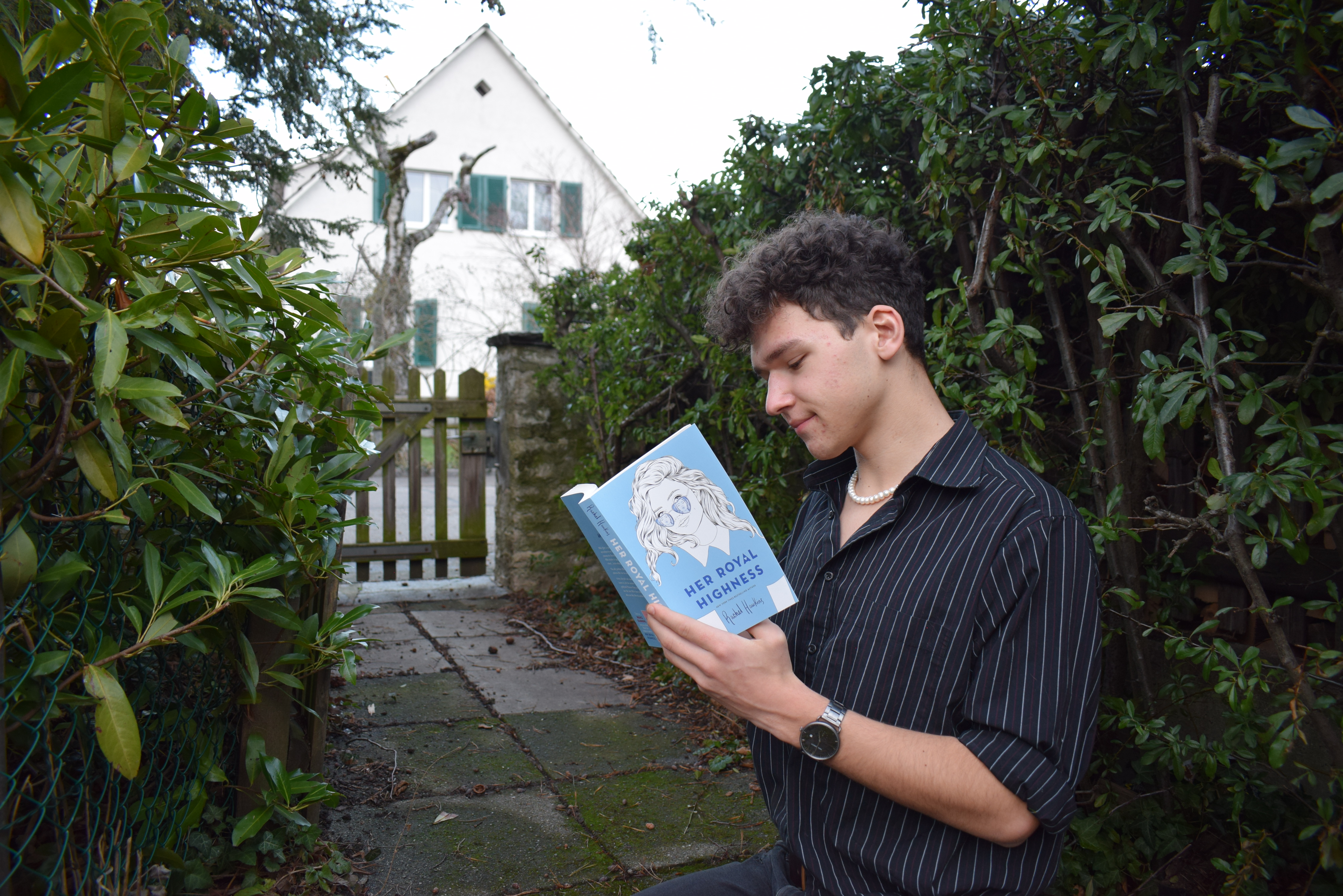 Royals: Herzensprinzessin - Her Royal Heighness Rezension - Josia Jourdan - young man with brwon curly hair and a dark blue shirt reading Her Royal Highness by Rachel Hawkins in front of a bush