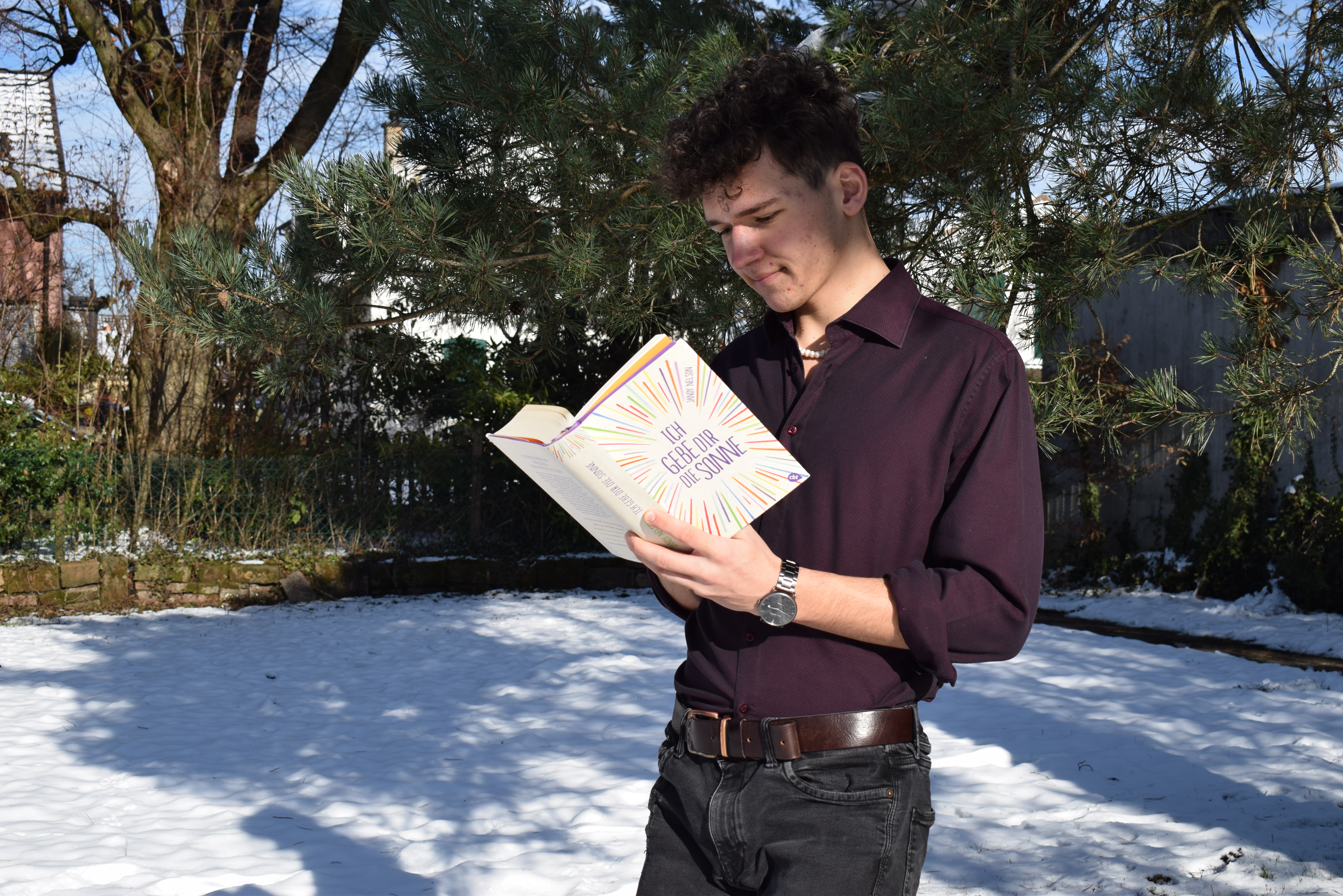 Junger Mann in violettem Hed im beschneiten Garten. er hält das Buch Ich gebe dir die Sonne von Jandy Nelson in den Händen.das Queere Repräsentation in Büchern - Josia Jourdan