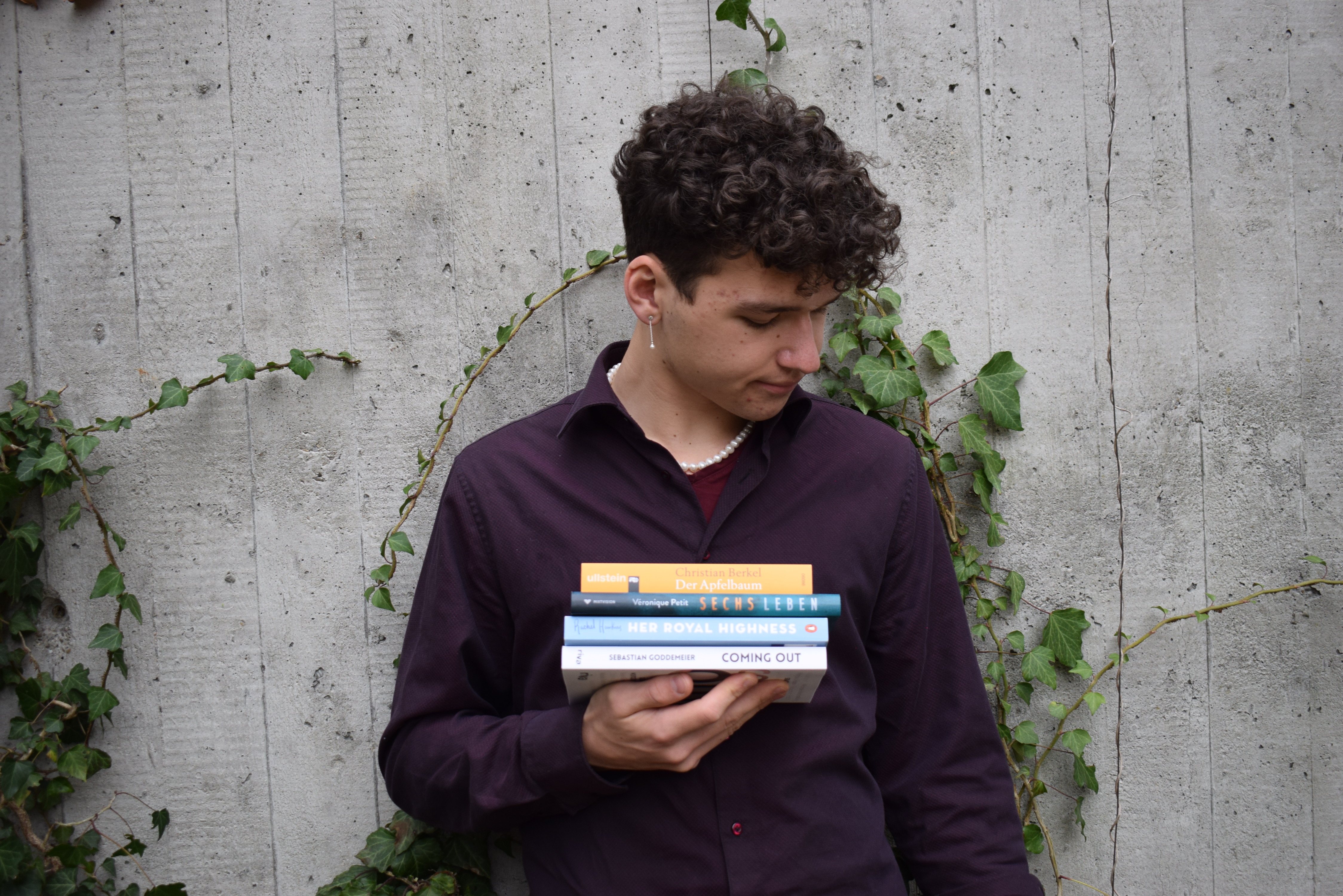 Monatsrückblick Februar 2021 - Josia Jourdan vor einer grauen Wand, violettes Hemd, Perlenkette und einen Bücherstapel in den Händen e